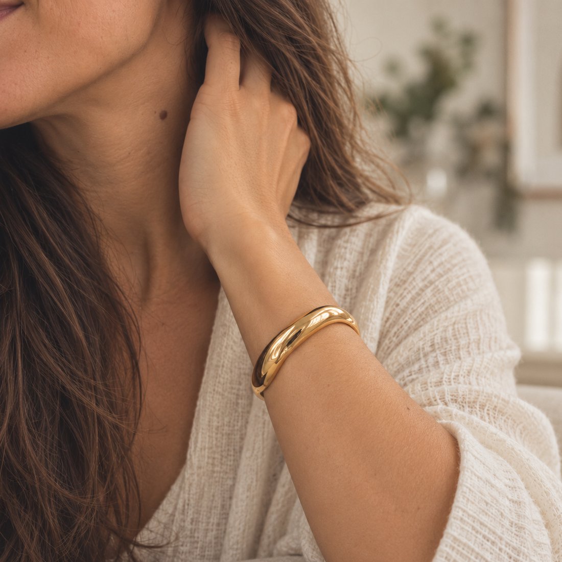 Pulseira rígida dourada no pulso, com a mão no cabelo.