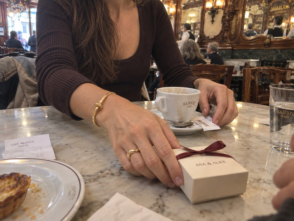 Mão pousada numa caixa de jóias bege com laço bordô, na mesa de um café, com uma pulseira dourada com nó.