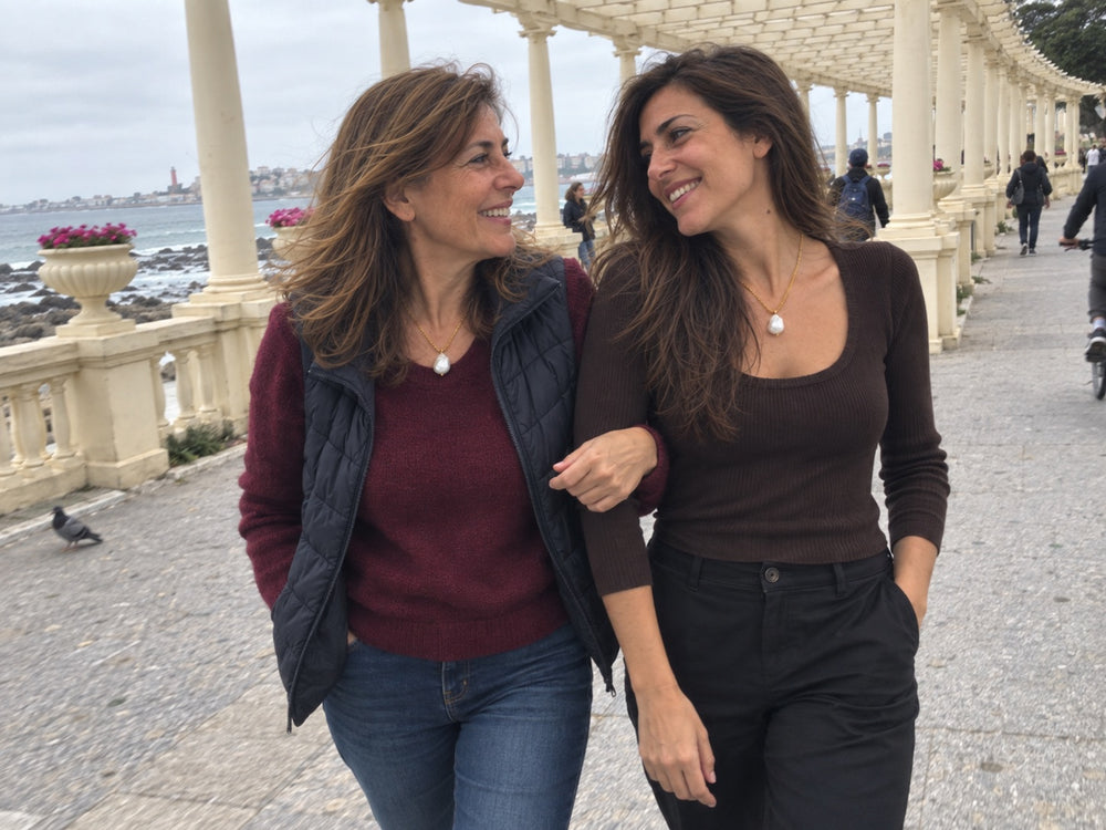 Mãe e filha de braço dado num passeio à beira-mar, as duas com o mesmo colar de pérola barroca.