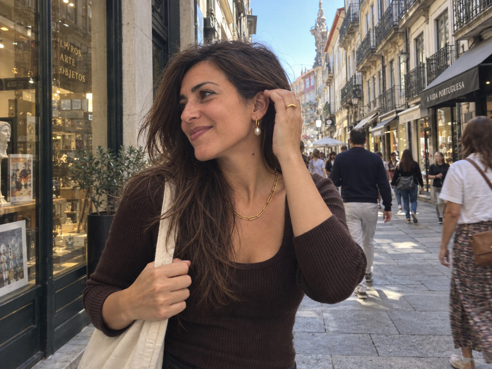 Mulher numa rua de lojas a ajeitar o cabelo, com argolas de pérola, colar de elos e anel dourados.