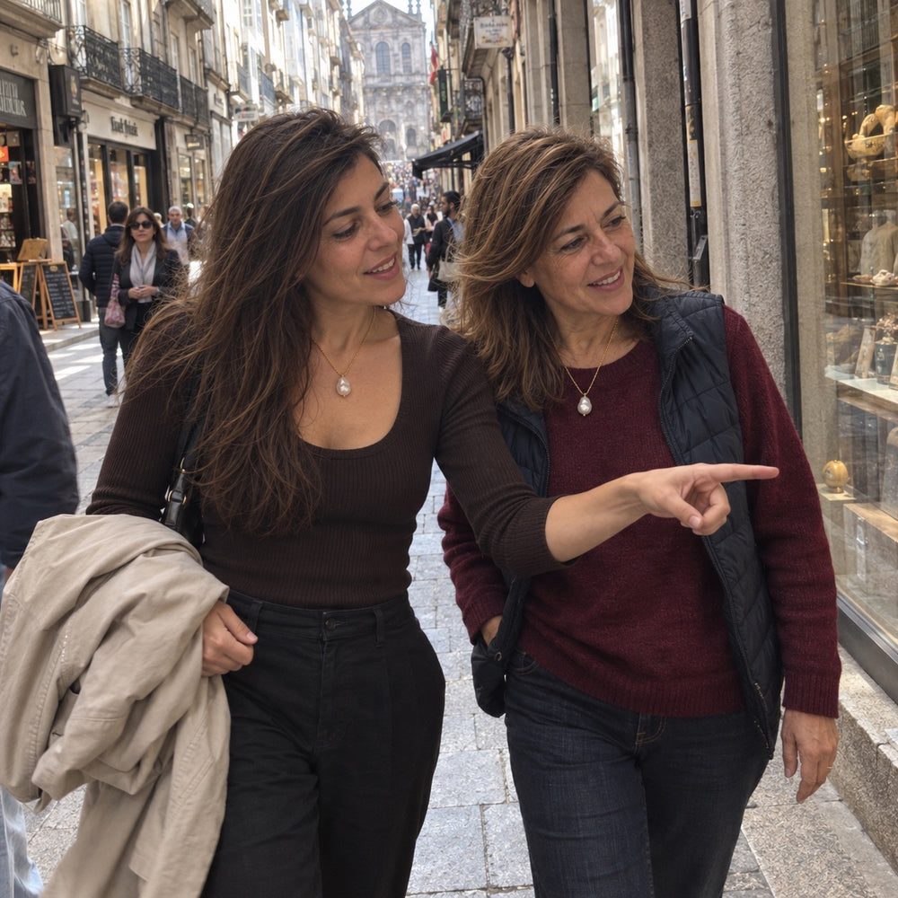 Mãe e filha a passear numa rua de lojas, com o mesmo colar de pérola barroca; a filha aponta para uma montra.